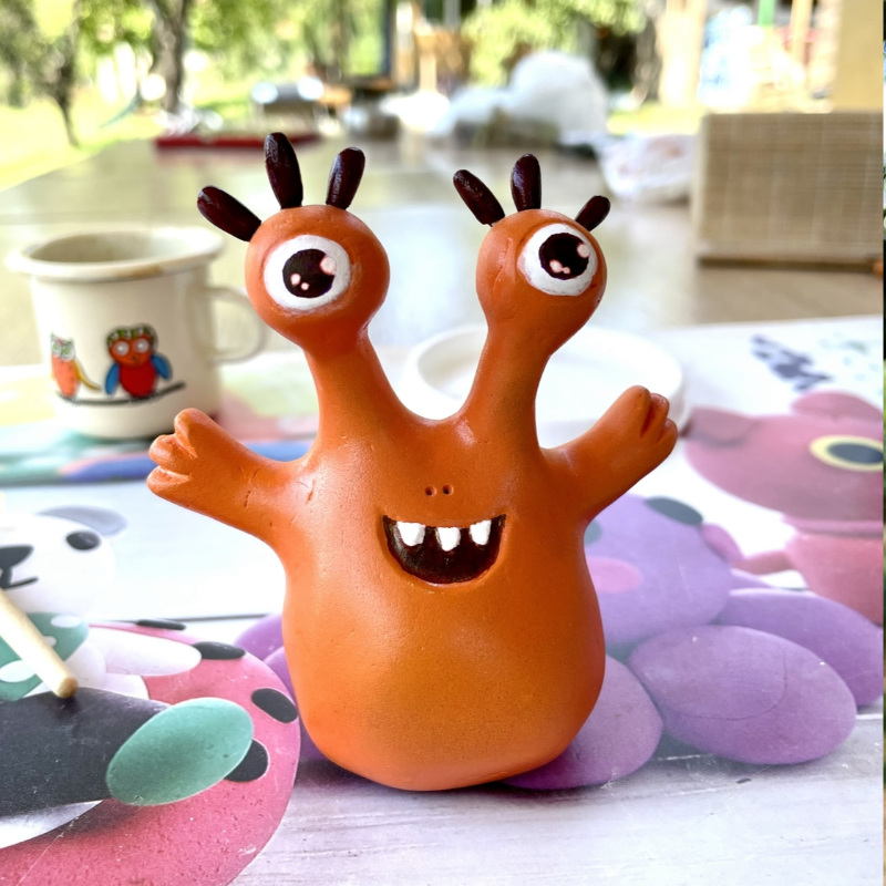 cute orange monster clay figurine with big eyes, fun eyelashes and a smile with big teeth showing with its hands wide open, ready for a hug. crafted by Boriana Giormova, Sofia, Bulgaria